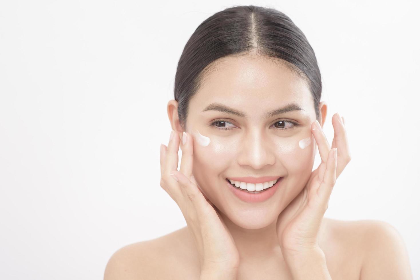 cerrar el retrato de belleza de una joven hermosa mujer sonriente aplicando crema facial sobre un estudio de fondo blanco foto