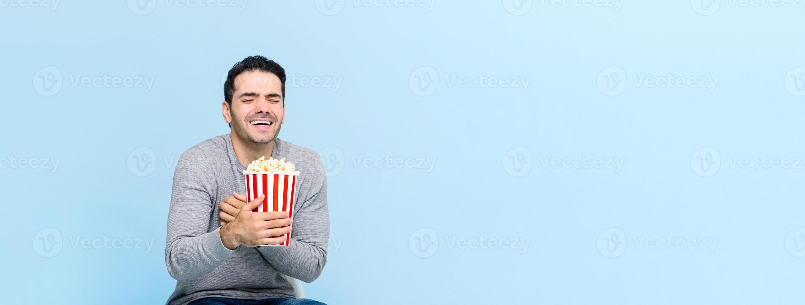 Young man holding popcorn laughing while watching movie isolated on light blue banner background with copy space photo