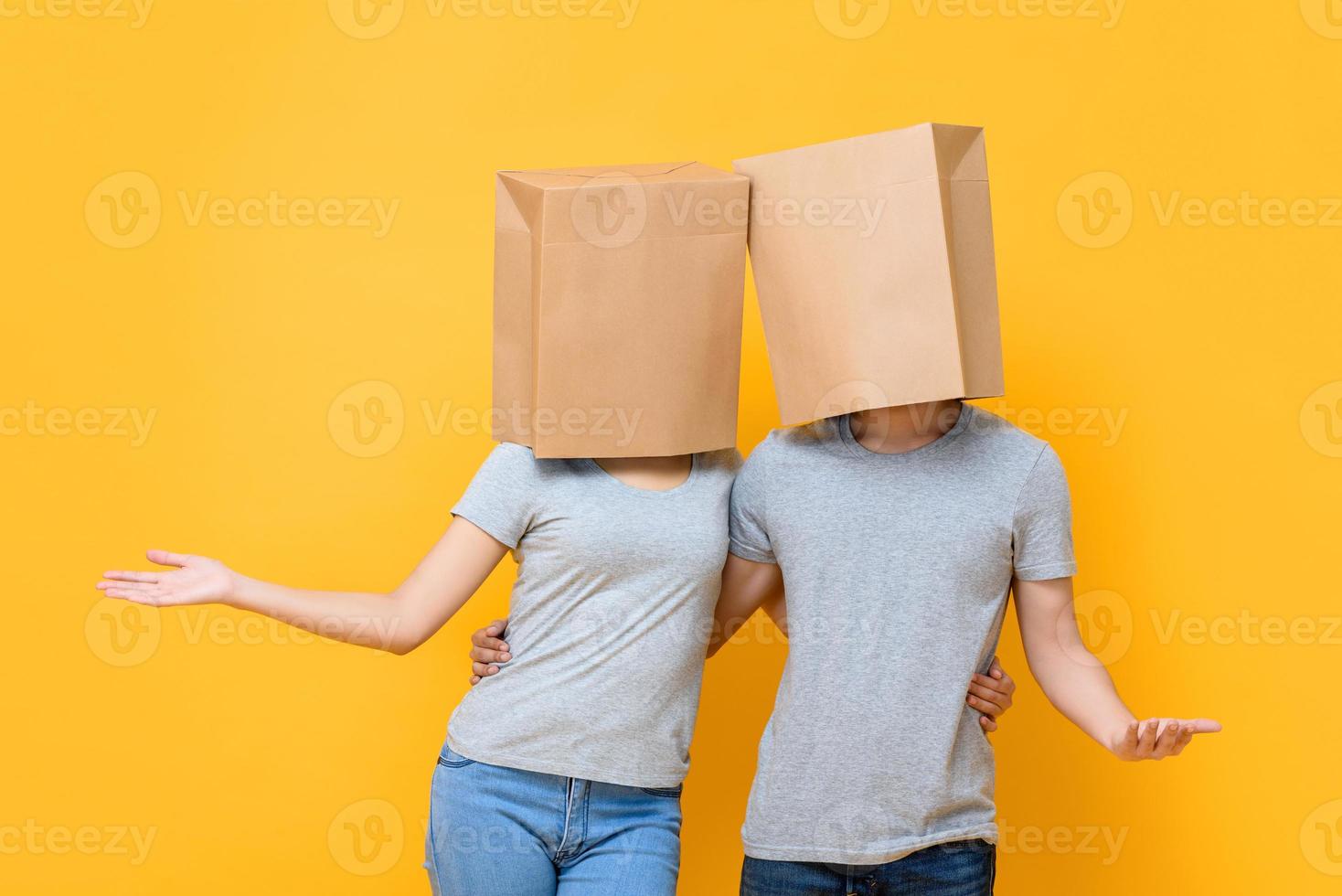 Faceless anonymous couple covering heads with paper bags and holding each other doing open hand gesture isolated on yellow studio background photo