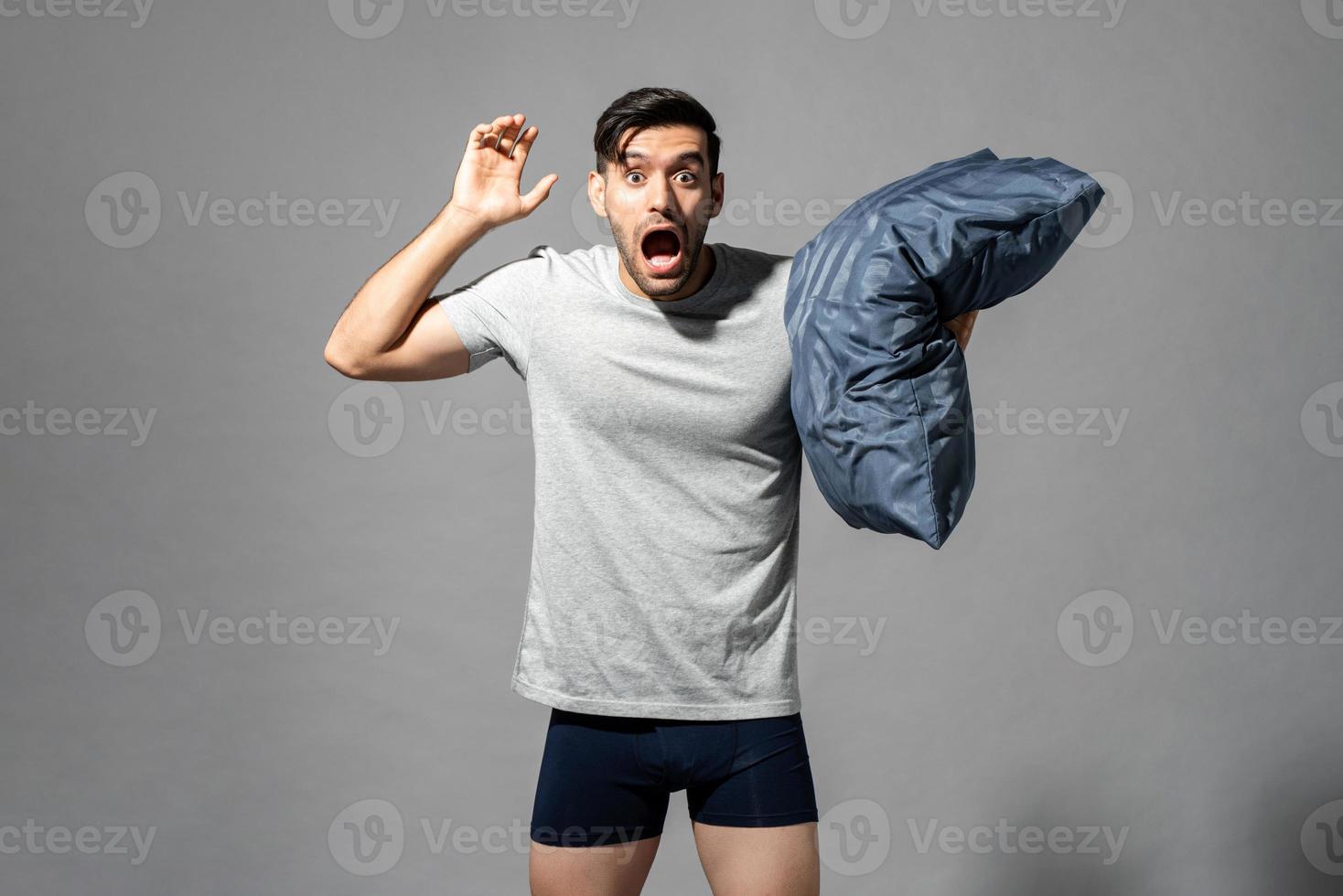 Sleepy young man in sleepwear feeling scared by nightmare and holding pillow, studio shot in isolated gray background photo