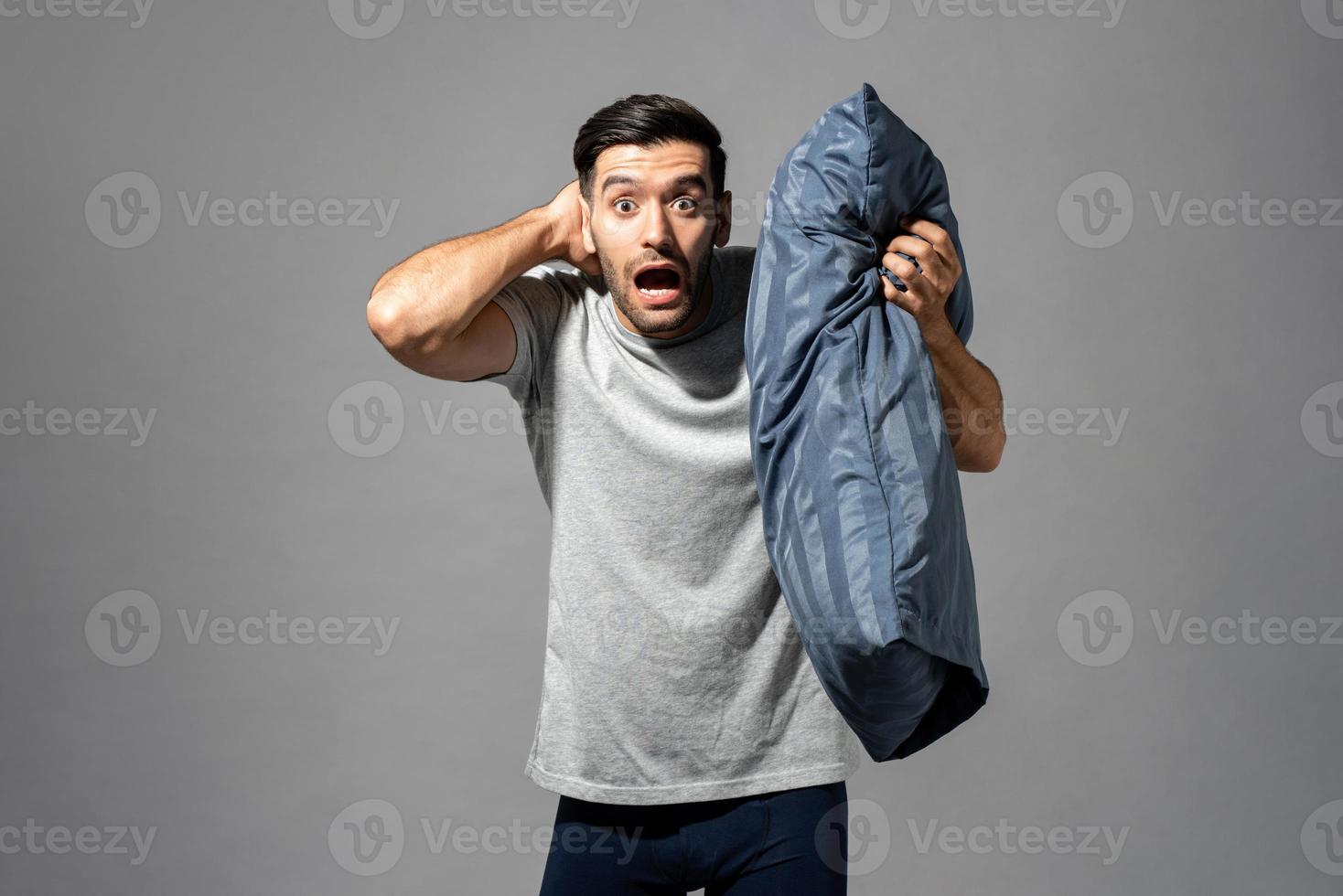 Young Caucasian man in sleepwear holding pillow waking up late and being shocked in light gray isolated studio background photo