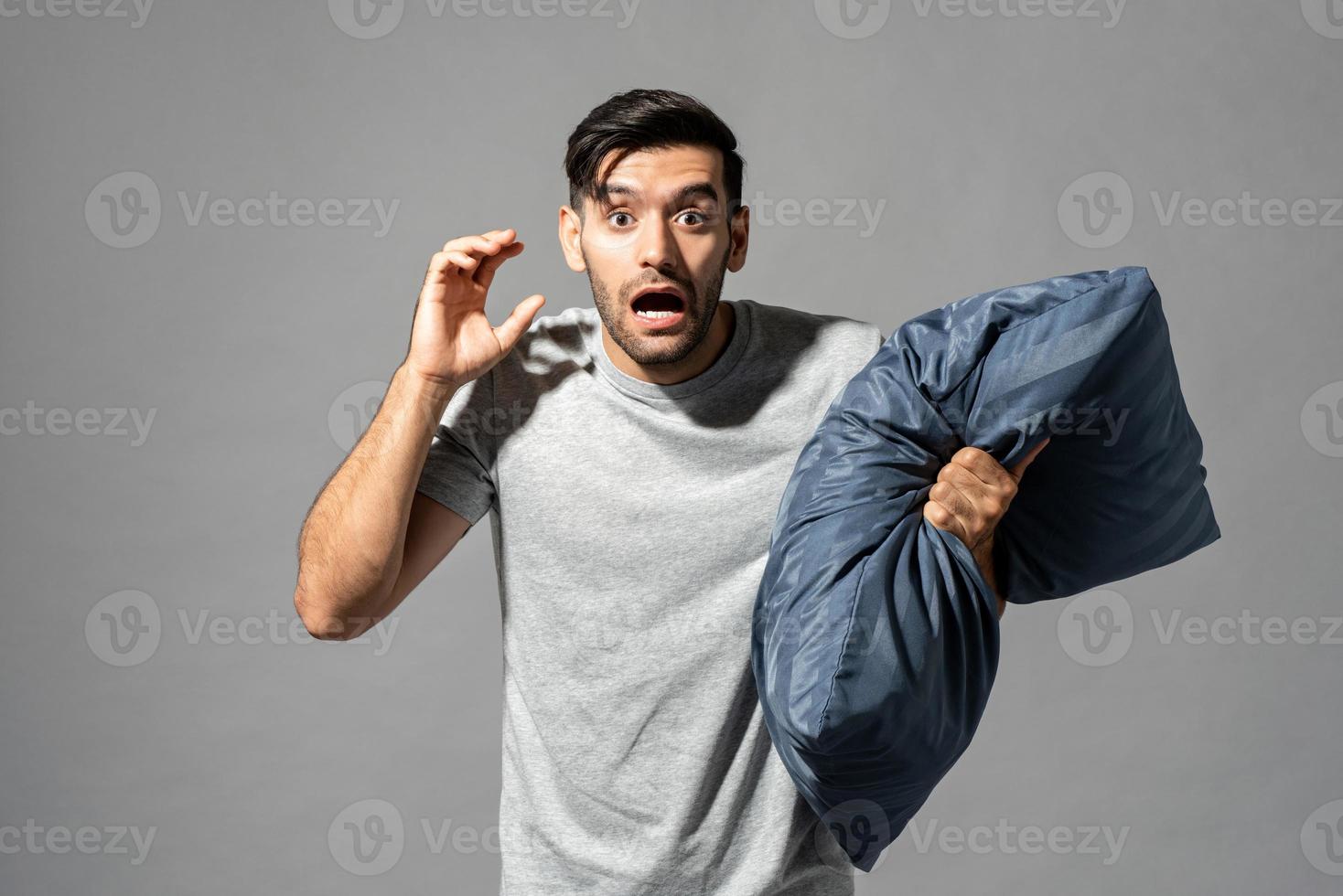 Sleepy young man in sleepwear feeling scared by nightmare and holding pillow, studio shot in isolated gray background photo