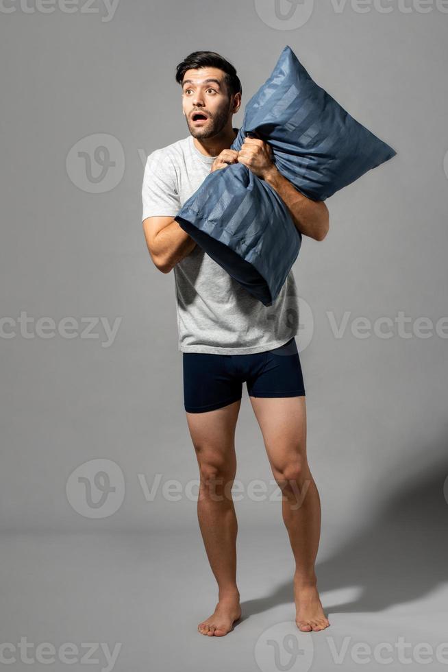 Full lenght portrait of young Caucasian man holding pillow and being shocked after waking up, studio shot in light gray isolated background photo