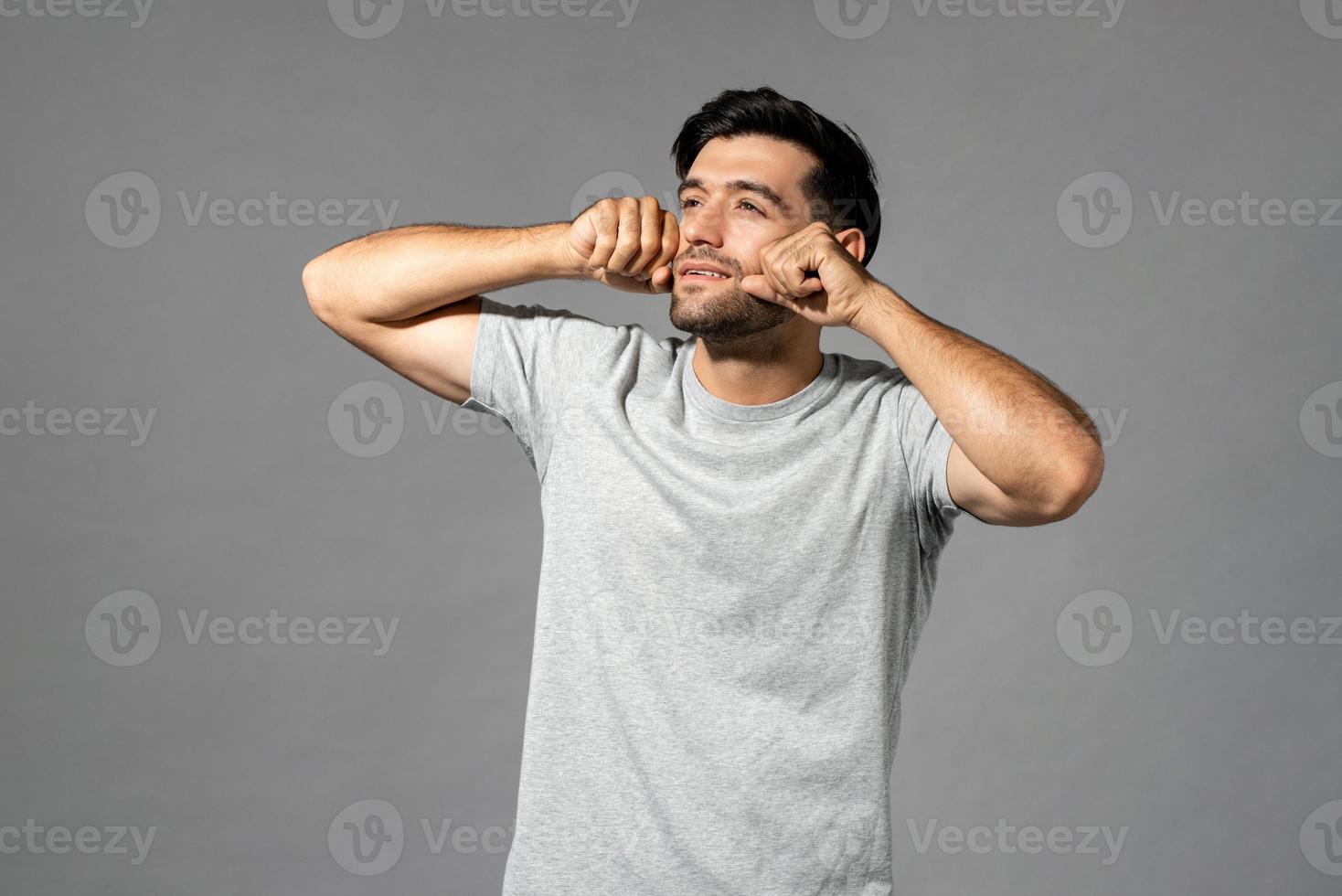 Portrait of sleepy young handsome Caucasian man rubbing eyes after wake up and looking away in isolated light gray studio background photo