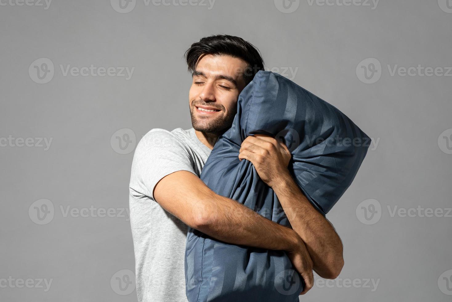 Sleepy young man holding pillow closing his eyes dreaming and smiling in isolated light gray studio background photo