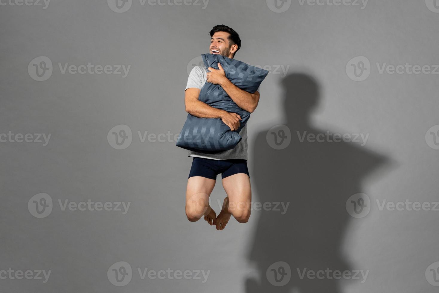 Portrait of young fresh energetic man wearing sleepwear holding pillow and jumping in mid-air after wake up from a good sleep in the morning,, studio shot in gray isolated background photo