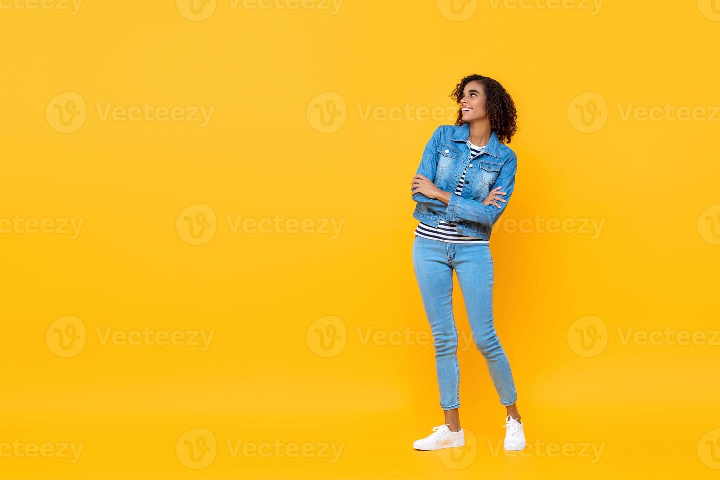retrato completo de una joven afroamericana sonriente mirando al costado con los brazos cruzados en el fondo amarillo del estudio foto