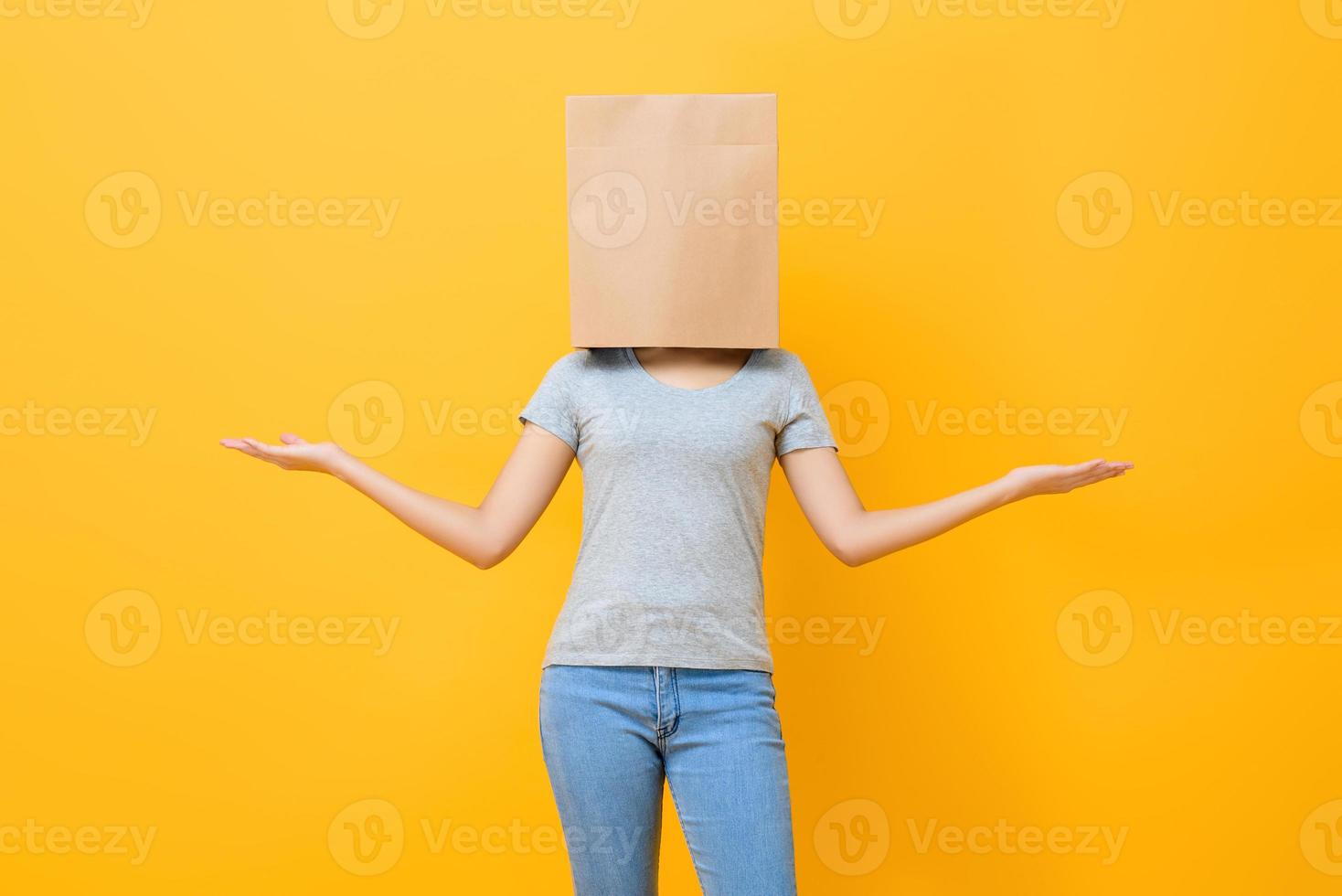 Anonymous woman in casual attire cover head with paper bag doing open hand gesture in yellow studio background photo