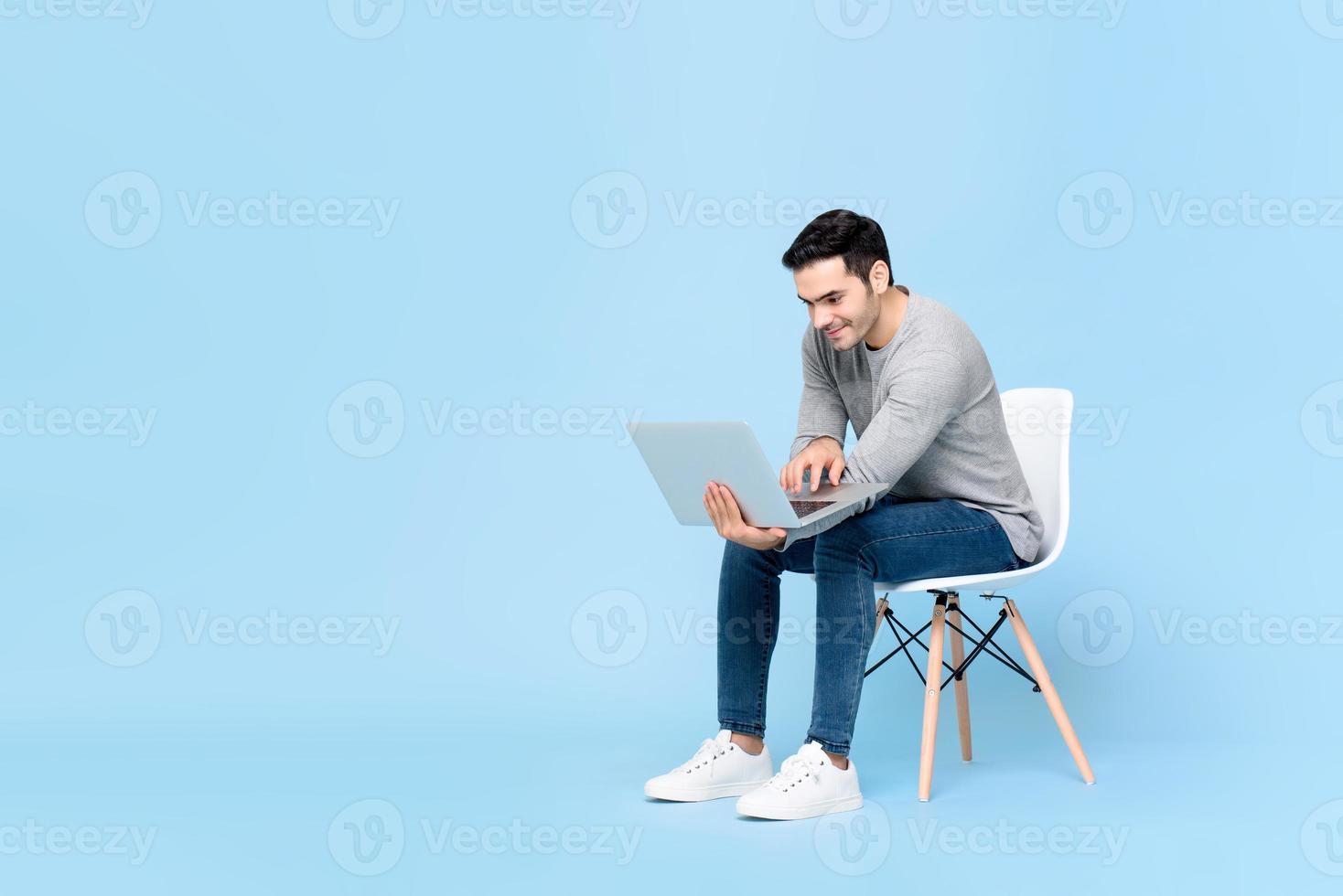 Portrait of smiling young handsome Caucasian man sitting while working on his laptop in isolated studio blue background photo