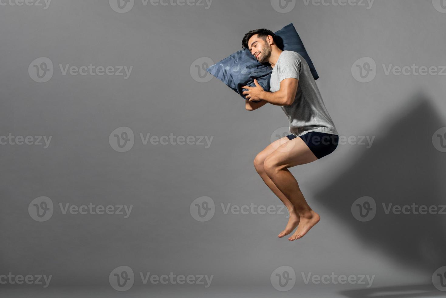 Young sleeping man holding his pillow levitating in empty gray room background with copy space photo