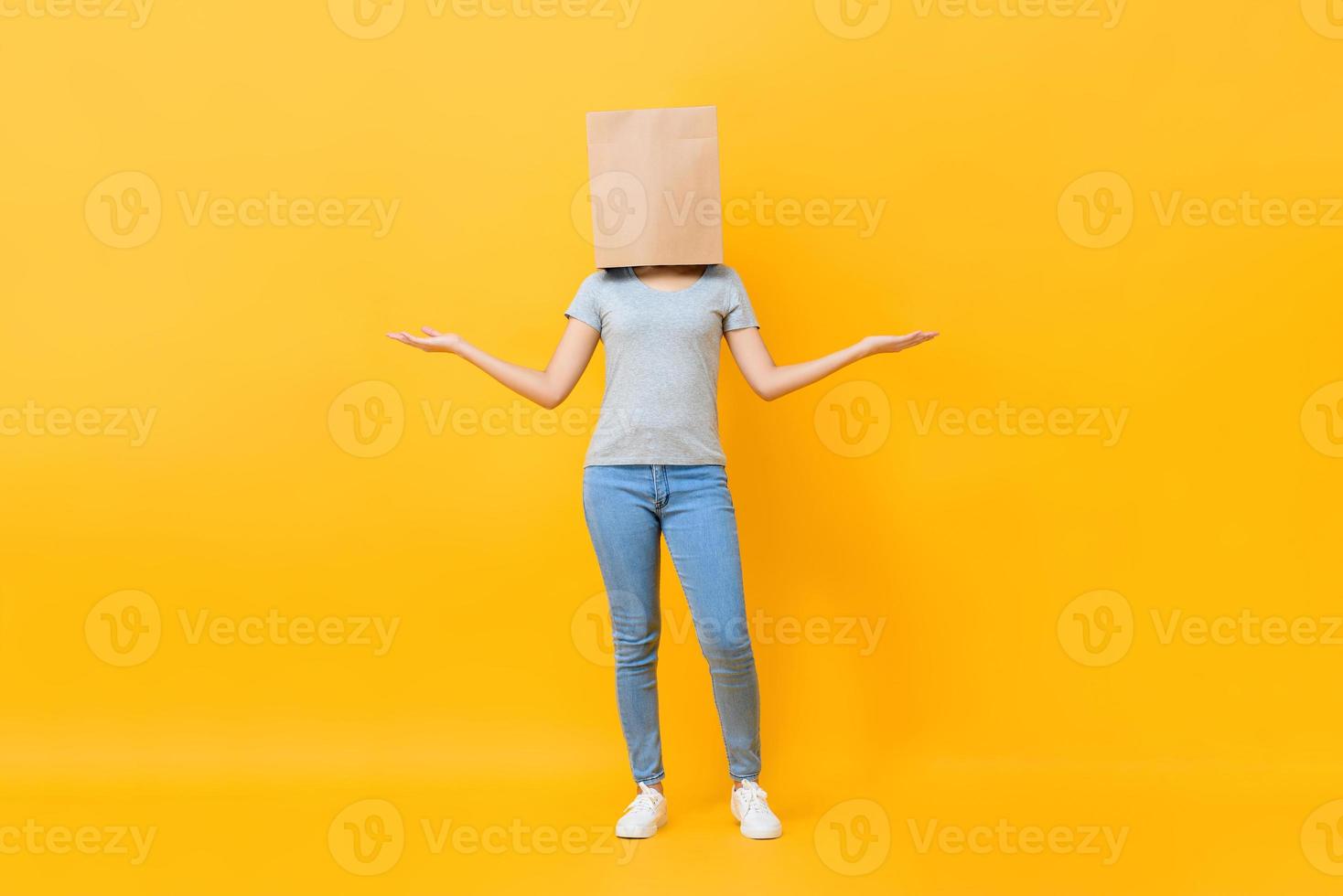 Anonymous woman in casual attire cover head with paper bag doing open hand gesture in yellow studio background photo