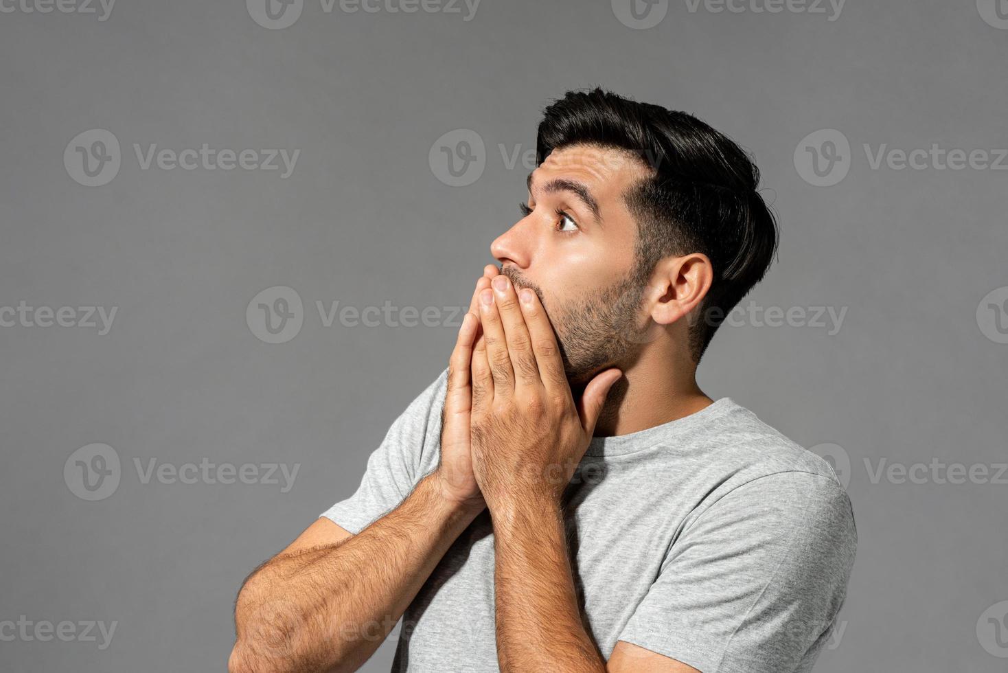 Shocked scared young Caucasian man with eyes popping and hands covering mouth on light gray studio background photo