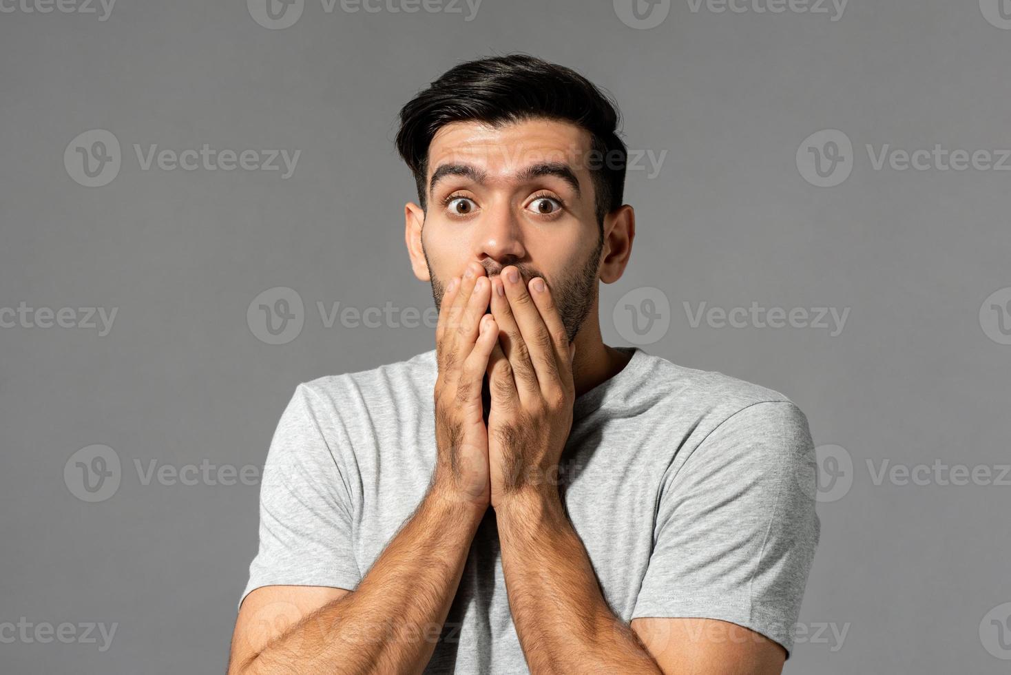 Shocked scared young Caucasian man with eyes popping and hands covering mouth on light gray studio background photo