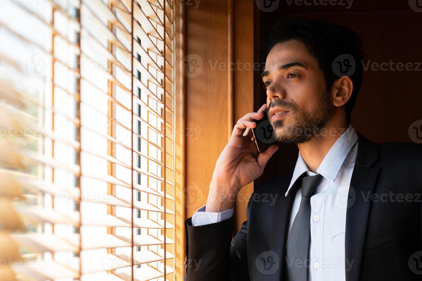 portrait of young businessman looking out of window and using smartphone to talk with client. business concept photo