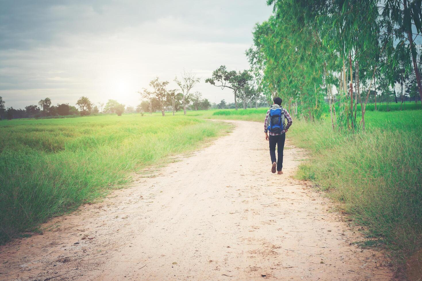 Back of young hipster man with backpack on his shoulder walking down countryside. photo