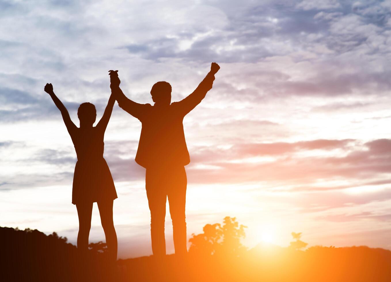 Silhouette of young couple together arms up in the air of happiness with accomplishment in the clouds at sunset. photo