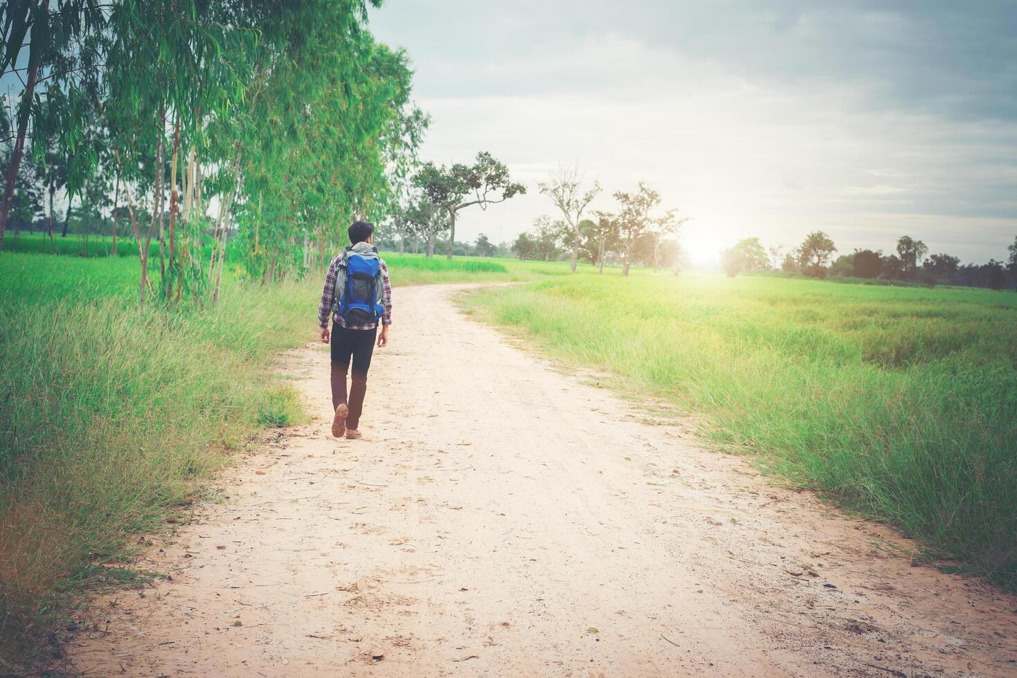 Back of young hipster man with backpack on his shoulder walking down countryside. photo