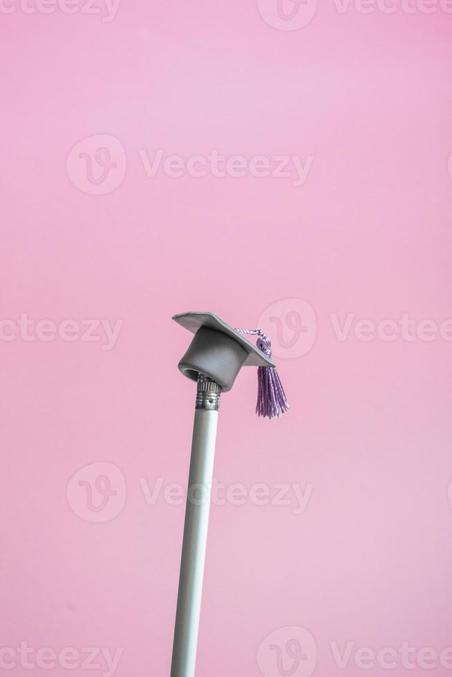 White pencil with graduation hat on a pink background closeup with copy