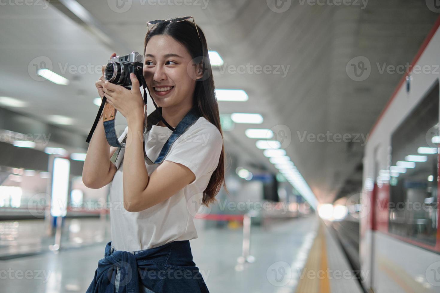 One young beautiful Asian female tourist, photographer taking snapshot photos with film camera, smile and enjoyment at train station platform, happy travel lifestyle by subway transport vacation trip.