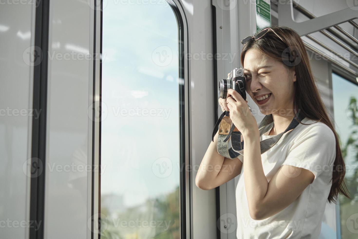 A Beautiful Asian female tourist with camera in the passenger cabin, traveling by sky train, taking snapshot photos when transporting in urban view, city lifestyle by railway, happy journey vacation.