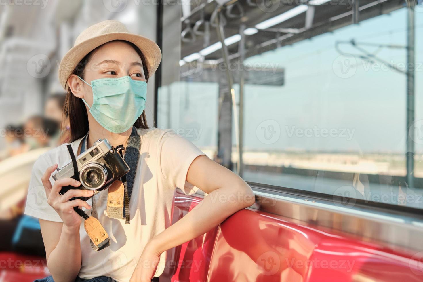 Beautiful Asian female tourist with face mask sits in a red seat, traveling by train, taking snapshot photo, transporting in suburb view, enjoy passenger lifestyle by railway, happy journey vacation. photo