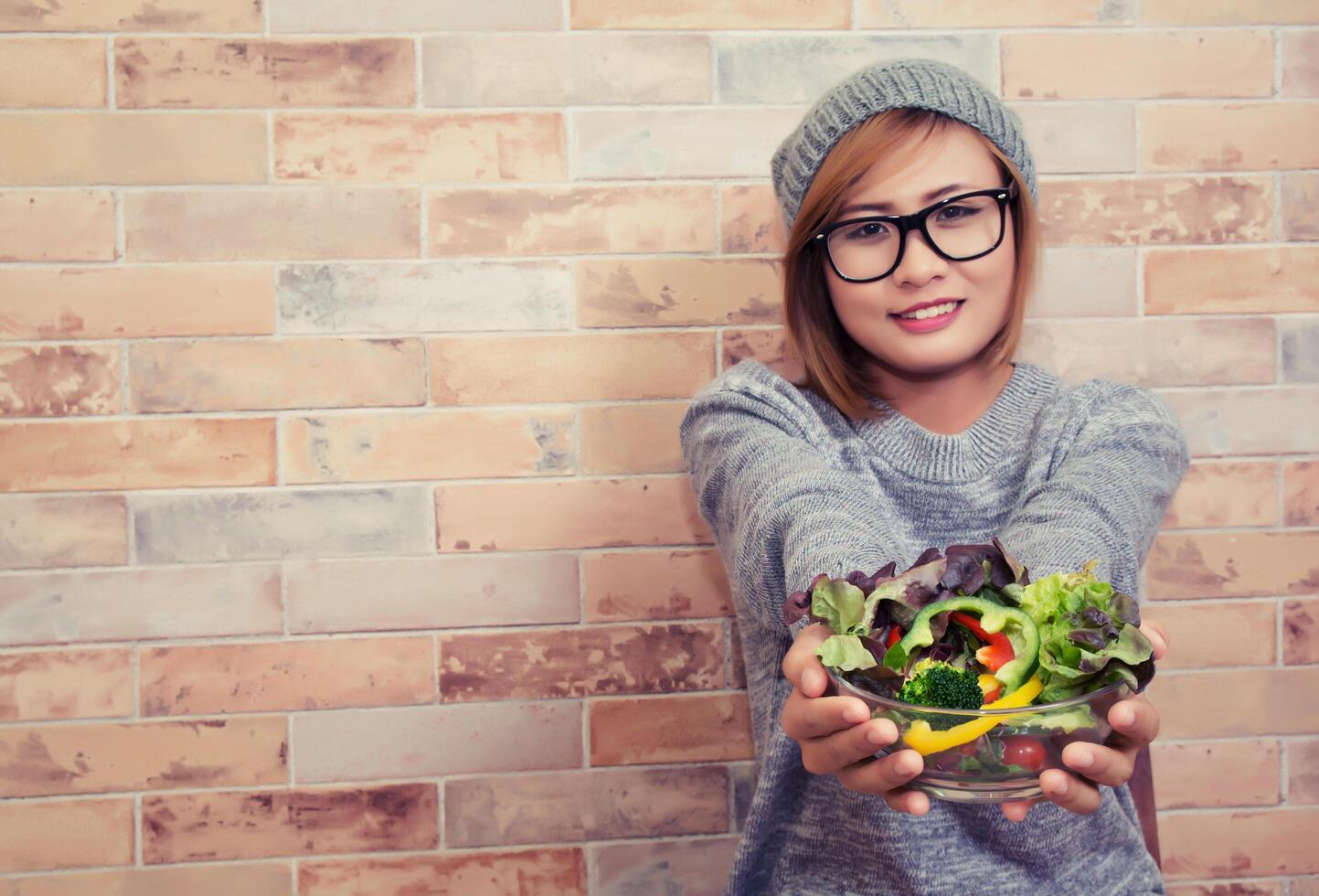 mujer hipster con gafas sentada en la silla con su ensalada sonriente a la cámara en el fondo de ladrillo foto