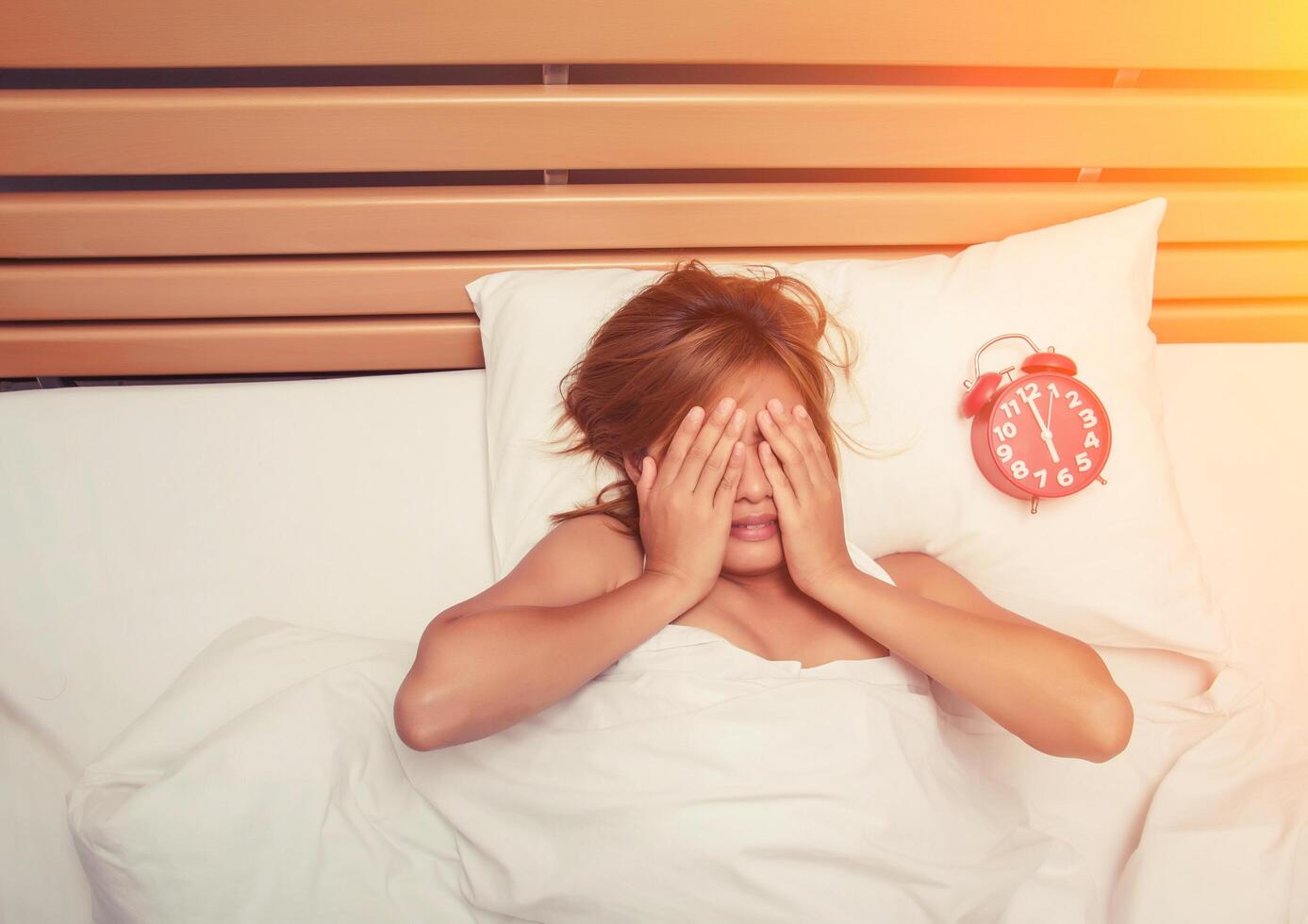 beautiful young woman take hands off her face on the bed in the morning when alarm clock is ringing don't want to wake up photo