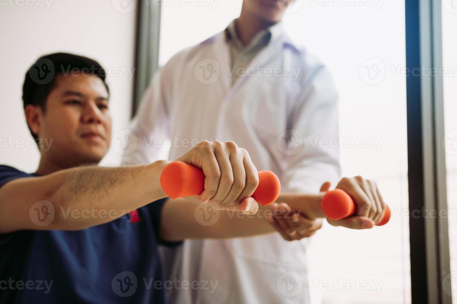 Fisioterapeuta masculino joven asiático que ayuda al paciente con ejercicios de levantamiento de pesas en el consultorio. foto