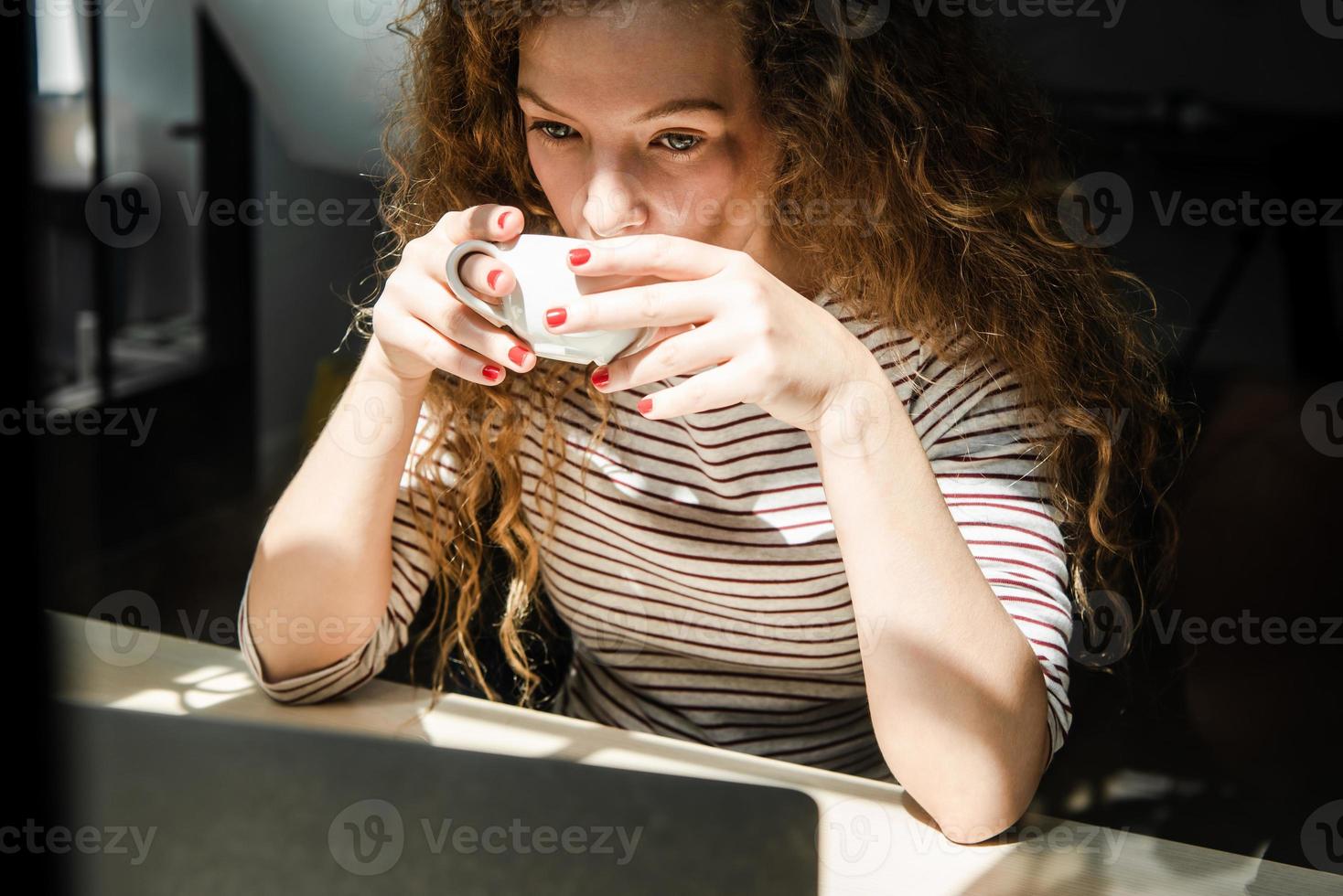 Young Caucasian woman drinking coffee while watching video on laptop computer at home in early morning photo