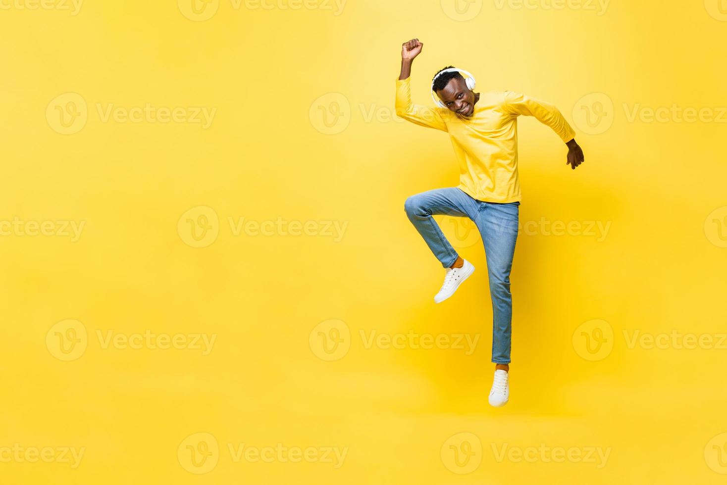 Happy energetic young African man wearing headphones listening to music and jumping with hand up next to empty space in yellow isolated studio background photo