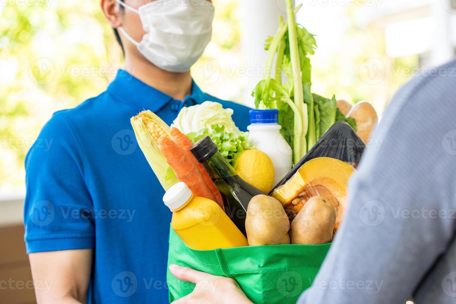 Asian deliveryman in blue uniform wearing face mask while delivering groceries to customer at home, food delivery in the time of pandemic concept photo