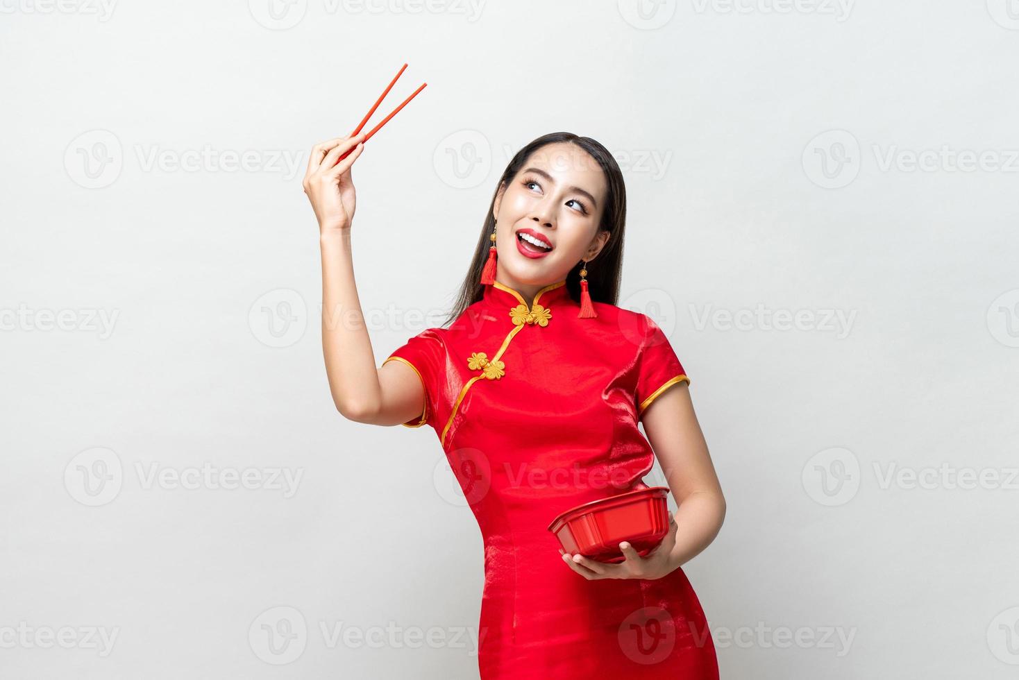 Cheerful Asian female in traditional red dress raising arm with chopsticks and looking up with smile against gray background photo