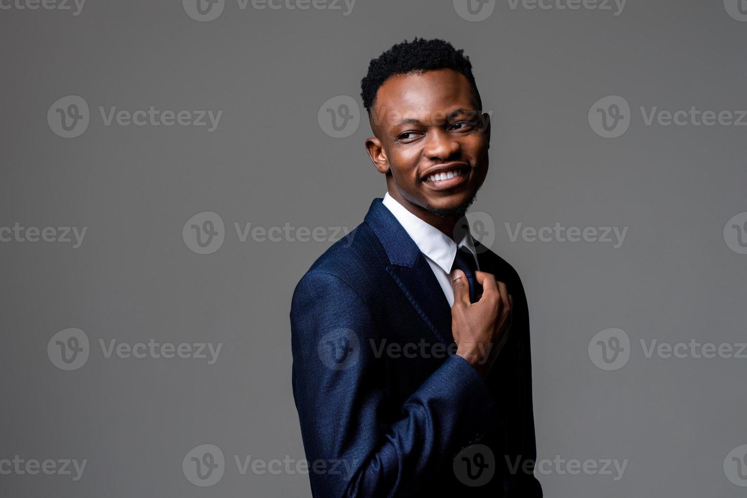 Smiling young handsome African gentle man wearing formal business suit looking aside with hand holding nectie isolated on studio gray background photo