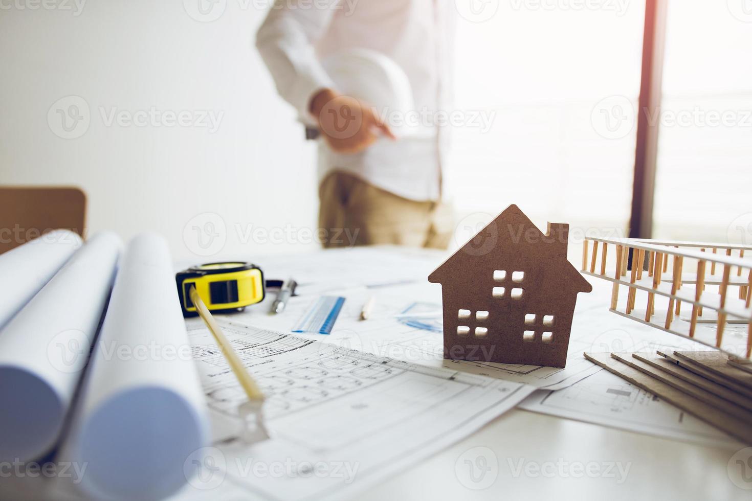 Engineer is holding a helmet standing in his office and has a blueprint and model on his desk. photo