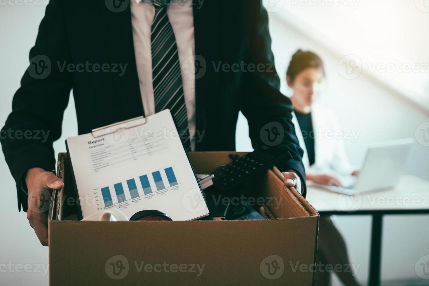 Front view with the male employee standing holding office supplies in the paper box going to submit a resignation letter while a female employee is working behind. photo