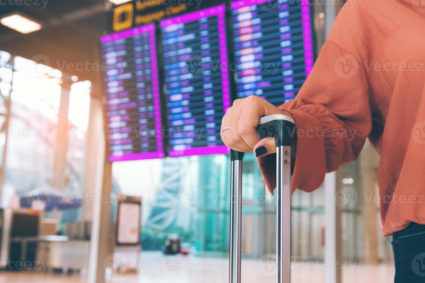 joven asiática parada en el aeropuerto sosteniendo una maleta revisando la salida con el horario de vuelo en segundo plano. foto