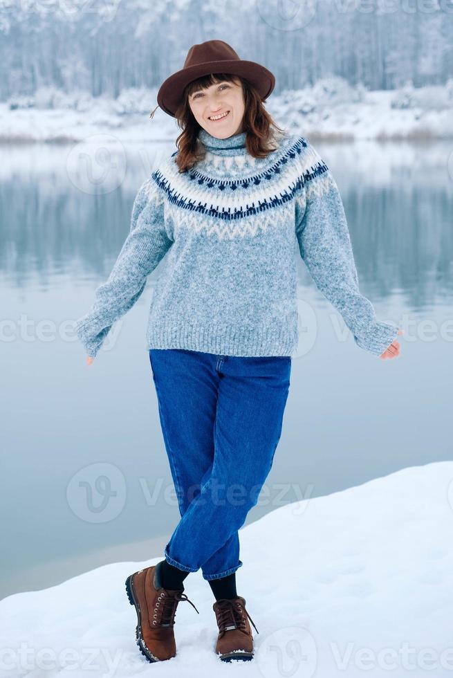 Smiling woman in a brown hat and sweater stands on the shores of a snow-covered lake and forest photo