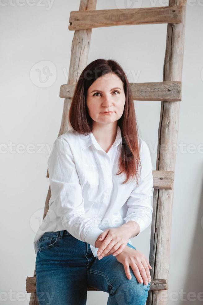 Beautiful woman dressed in a white shirt and blue jeans posing near a wooden ladder on a white background. Copy, empty space for text photo