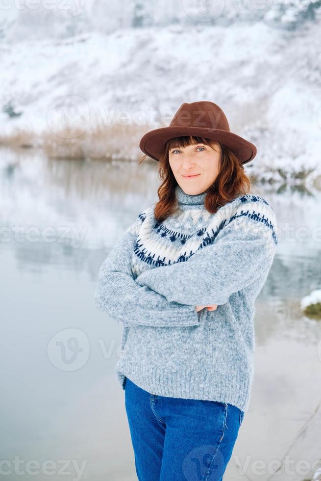 Woman in a brown hat and sweater stands on the shores of a snow-covered lake and forest photo