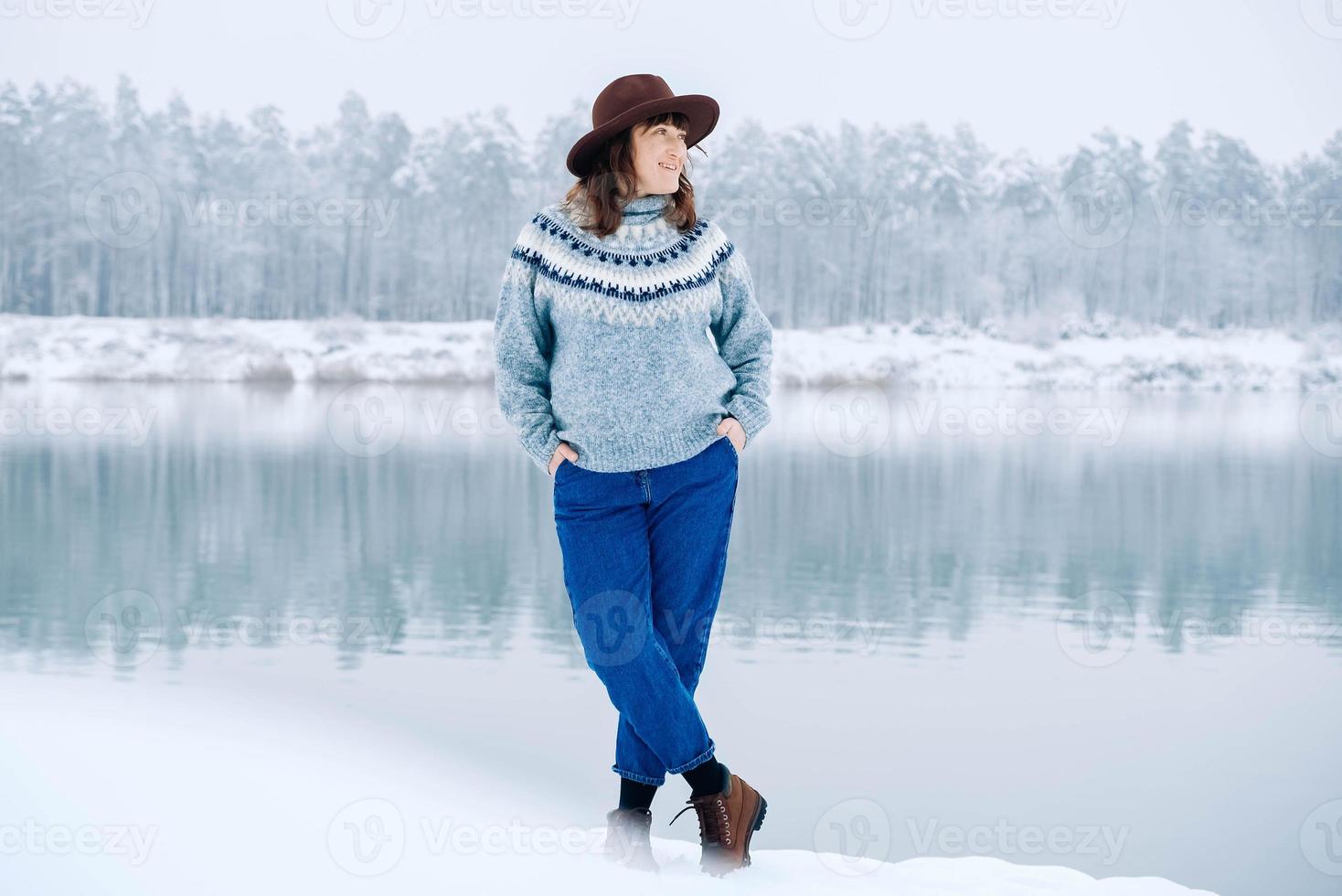 Smiling woman in a brown hat and sweater stands on the shores of a snow-covered lake and forest photo