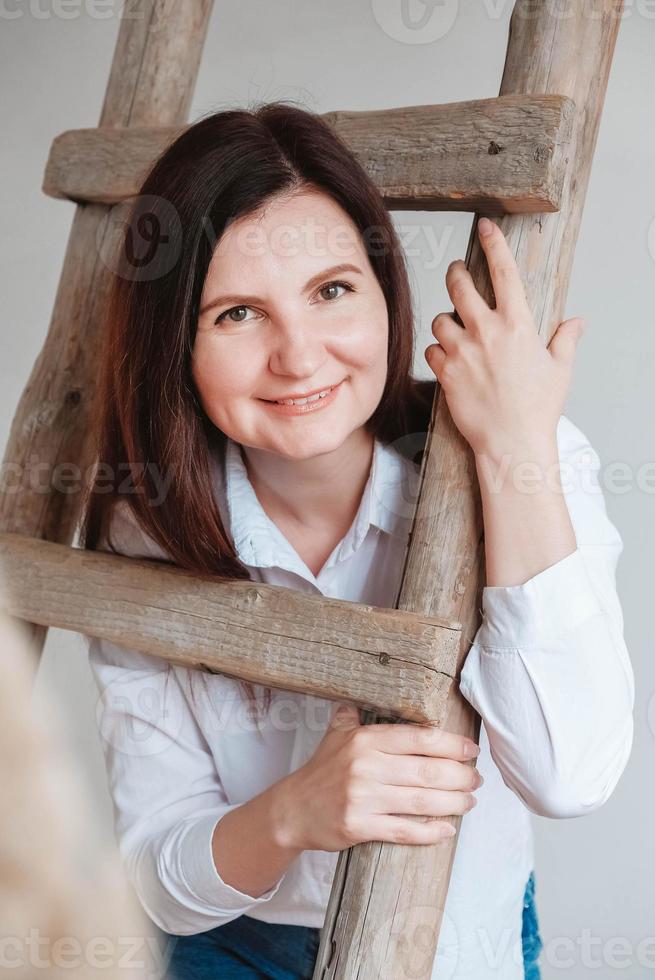 Beautiful woman dressed in a white shirt and blue jeans posing near a wooden ladder on a white background photo