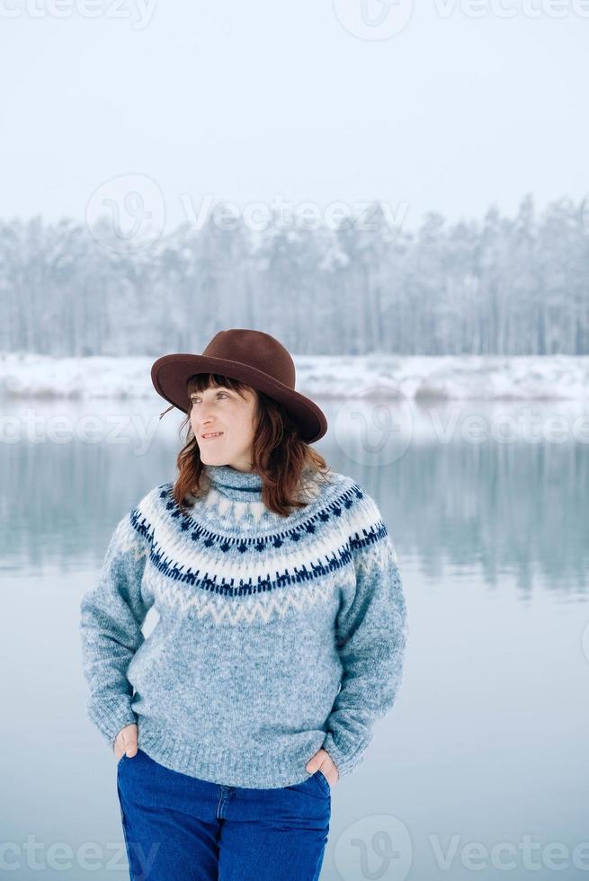 Woman in a brown hat and sweater stands on the shores of a snow-covered lake and forest photo