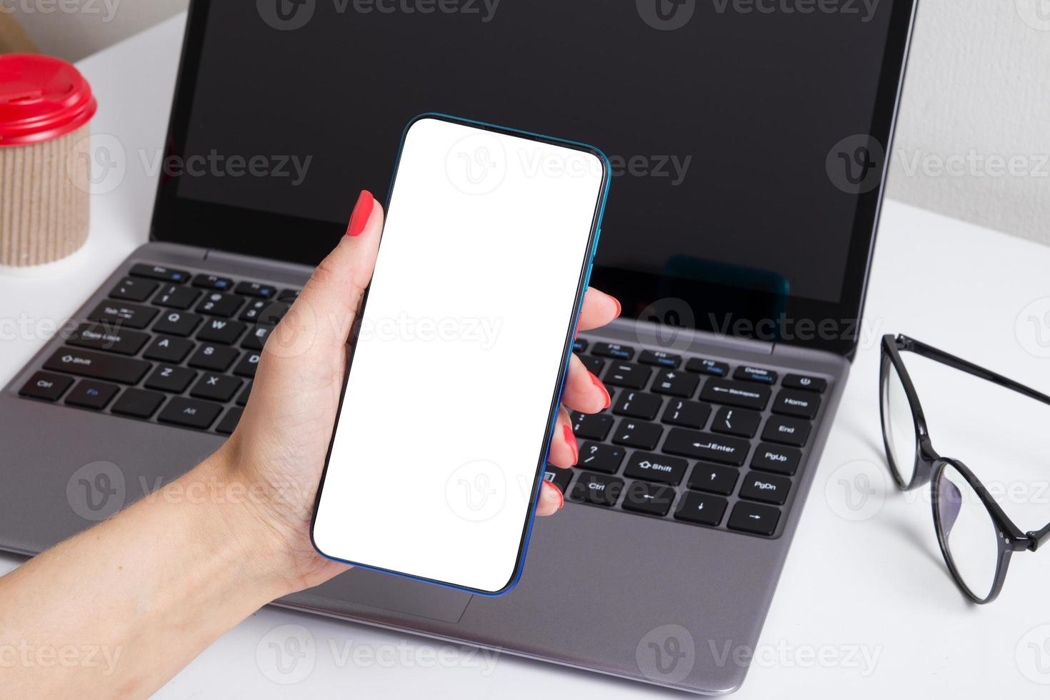 Phone in a female hand mock-up on the background of a laptop and coffee. Online shopping from home concept photo