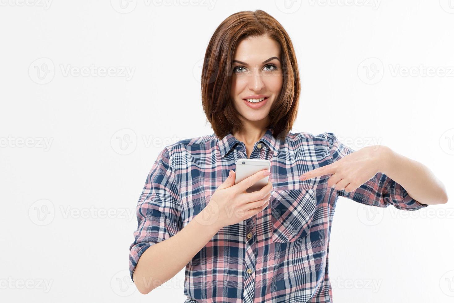 la niña sonriente está apuntando al teléfono inteligente de pie sobre fondo blanco. feliz y hermosa mujer joven sosteniendo el teléfono móvil y señalando con el dedo. foto