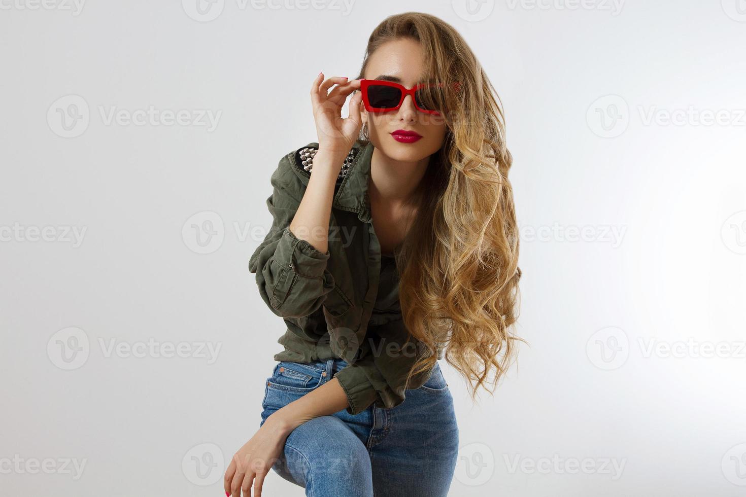 Stylish woman in red glasses looking at camera at studio isolated on white background. Copy space. Trendy style of beautiful luxury model with red lips makeup. Long wavy shiny blonde hair photo