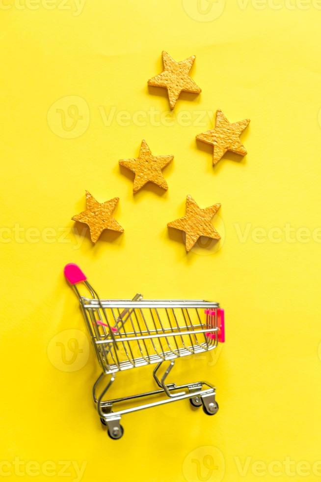 Simply flat lay design small supermarket grocery push cart for shopping with wheels and 5 gold stars rating isolated on yellow background. Retail consumer buying online assessment and review concept. photo