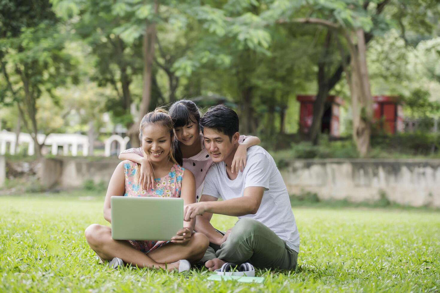 Happy family father, mother and daughter sitting on the grass and playing laptop at outdoor park photo