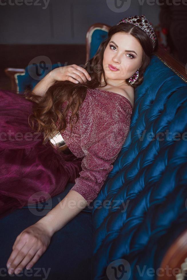 portrait of beautiful young pretty cute woman princess  in long purple queen's dress and crown, with long hair and make up sitting on the blue velvet sofa indoor in minimalistic interior photo