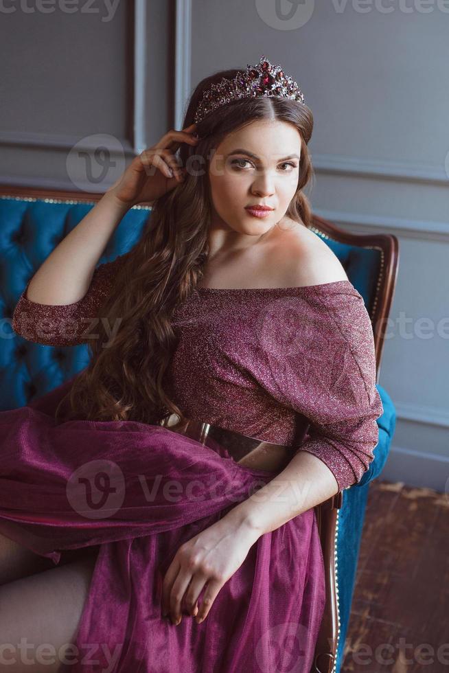 portrait of beautiful young pretty cute woman princess  in long purple queen's dress and crown, with long hair and make up sitting on the blue velvet sofa indoor in minimalistic interior photo