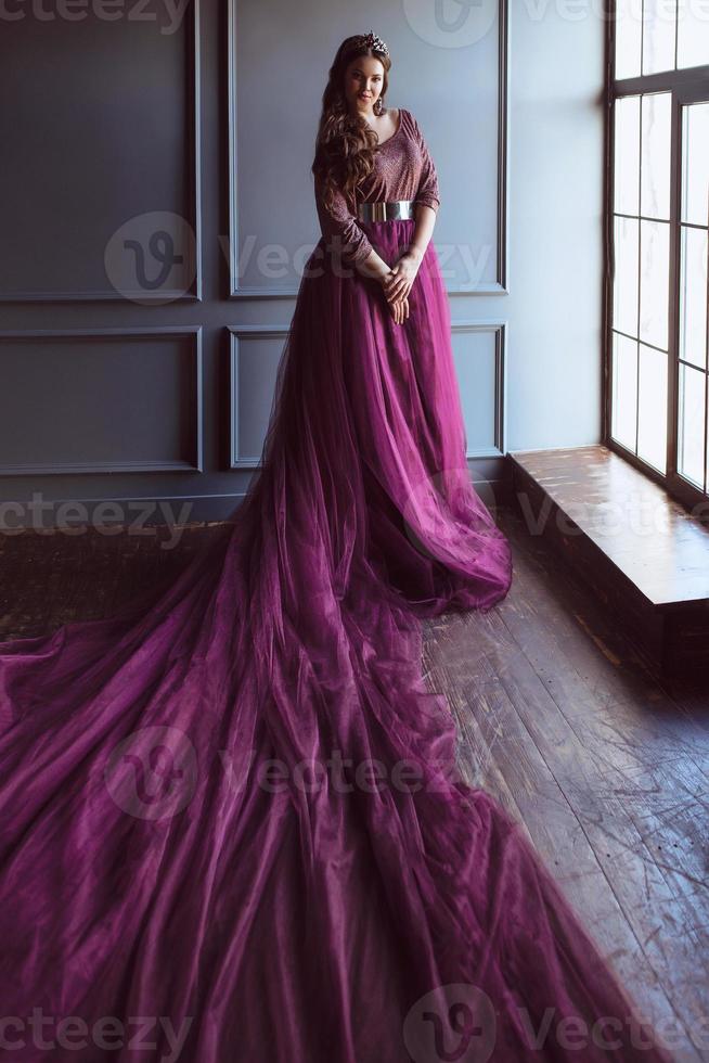 retrato de una hermosa joven princesa linda con un largo vestido de reina púrpura y una corona, con el pelo largo y maquillada en el interior de un loft minimalista de pie junto a la ventana foto